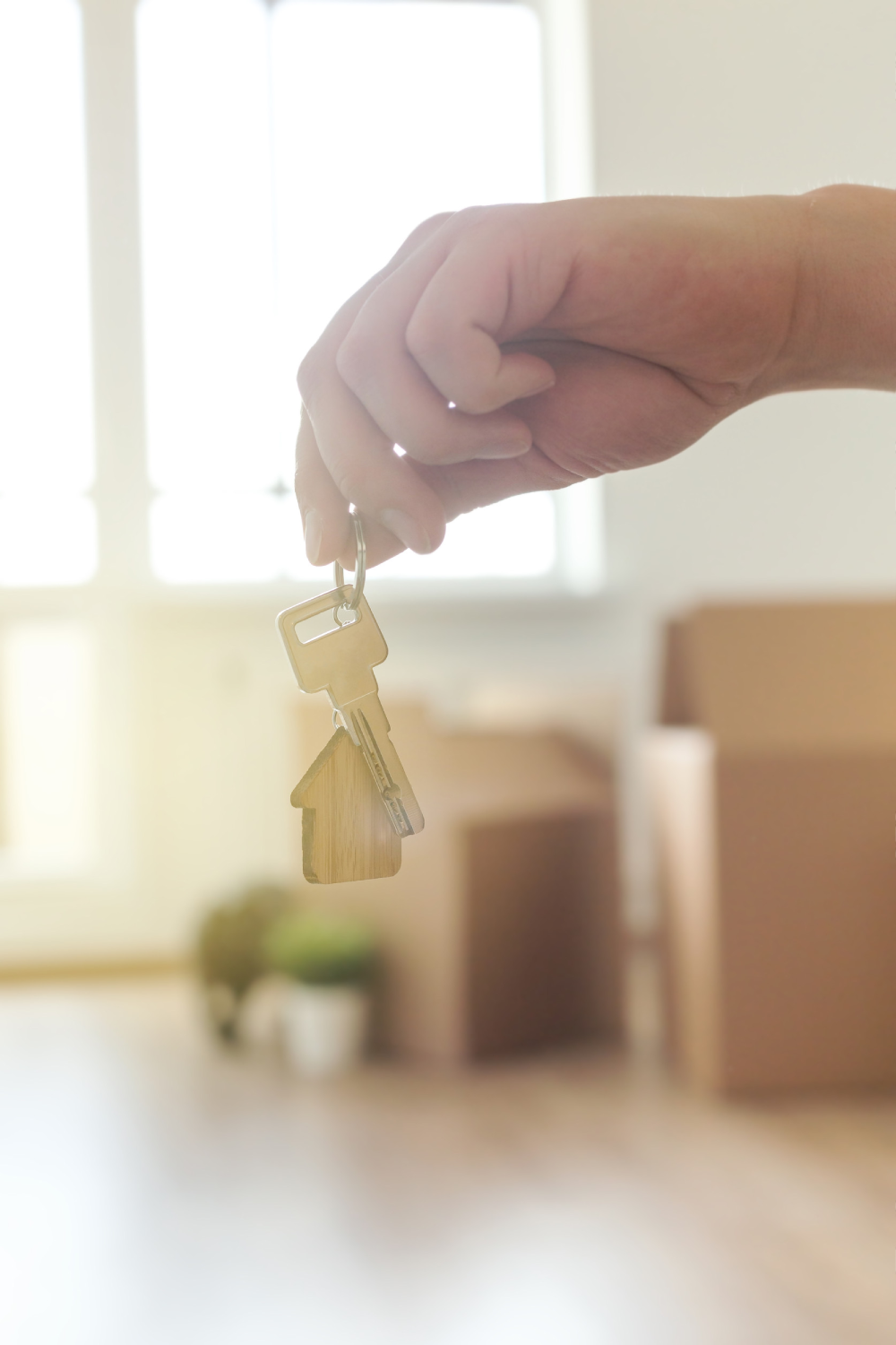 Home key in male hand on room background inside new house with cardboard boxes against the window. Real estate and moving and rent apartment concept